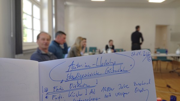 Eine Seite in einem Notizbuch mit der Überschrift '"Autor:innen-Workshop "Stadtgeschichte Gütersloh"', im Hintergrund ein Seminarraum mit Menschen