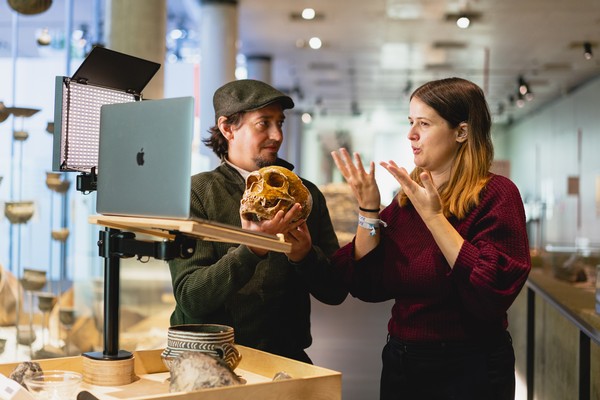 Zwei Personen stehen in einem Museum, eine hält einen Schädel, die andere gebärdet, im Vordergrund ein Laptop auf einem Ständer.