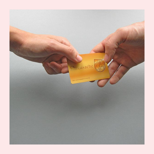 Zwei Hände übergeben eine goldfarbene Karte mit dem Schriftzug 'Ehrensache' und einem Wappensymbol. Foto eingerahmt mit rosa Rahmen.