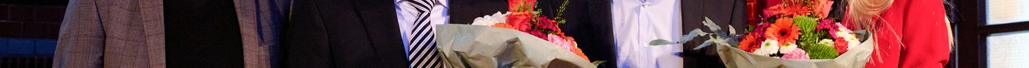 Vier Personen stehen nebeneinander und halten Blumensträuße sowie kleine Auszeichnungen in einem festlich beleuchteten Innenraum.