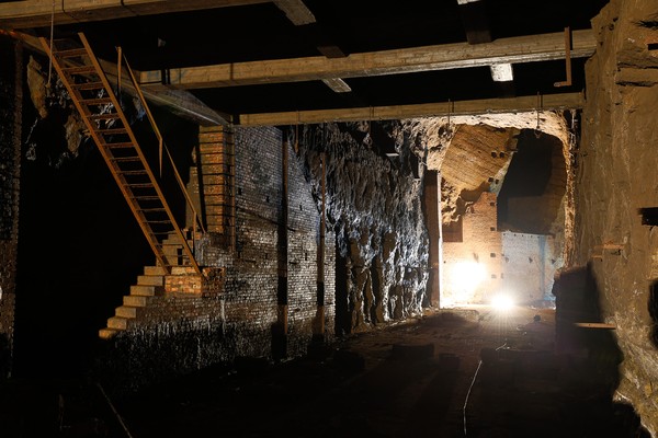 Unterirdische Struktur mit rauen Felswänden und Ziegelmauerwerk. Eine Metalltreppe führt zu einer höheren Ebene.