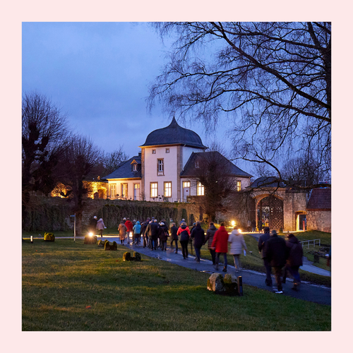Gruppe von Menschen geht auf beleuchteten Weg zu einem historischen Gebäude mit gewölbtem Dach bei Dämmerung. Foto eingerahmt von rosa Rahmen.