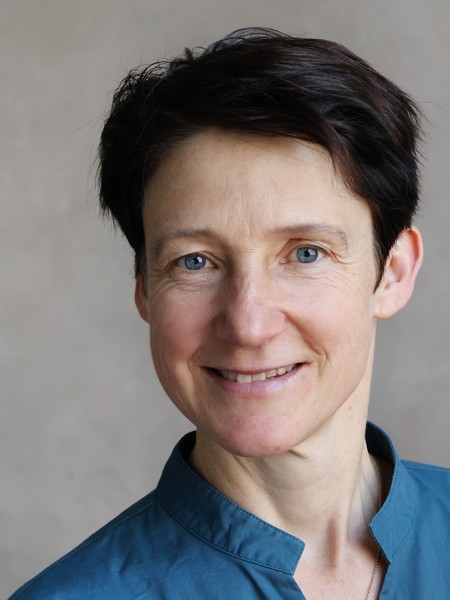 Frau mit kurzen dunklen Haaren und blauem Hemd vor neutralem Hintergrund lächelnd