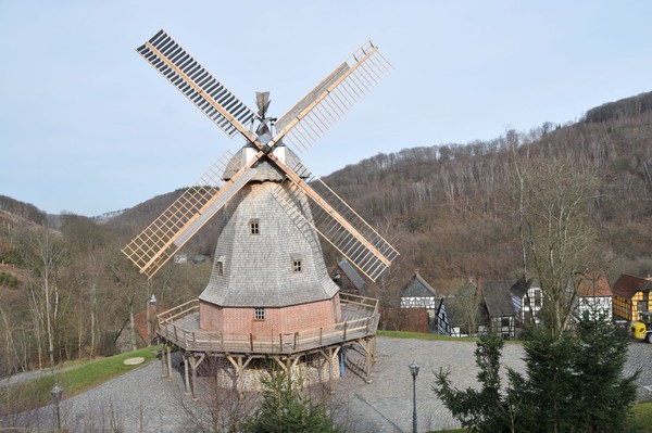 Historische Windmühle mit Holzflügeln auf rundem Steinsockel in ländlicher Umgebung mit Hügeln und Fachwerkhäusern im Hintergrund