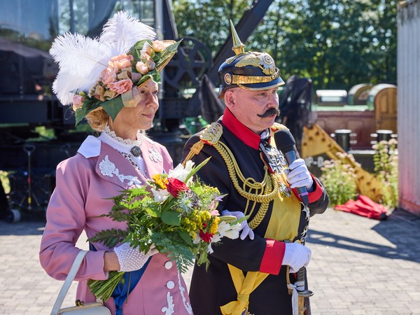Zwei Personen in Steampunk-Kostümen, eine Person trägt ein Kleid mit Spitze und einen Hut mit Federn, die andere eine Uniform mit goldenen Verzierungen, Pickelhaube und hält ein Mikrofon