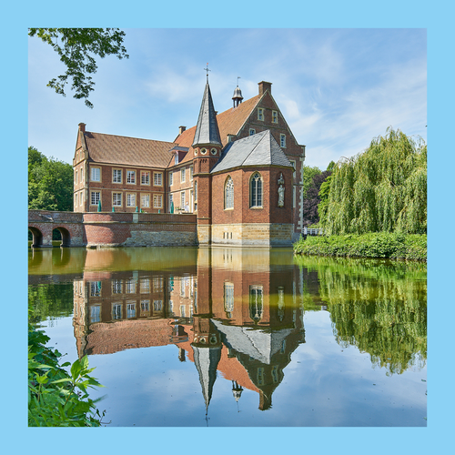 Foto eingefasst in blauen Rahmen. Foto von Burg umgeben von Wasser. Im Wasser des Burggrabens spiegelt sich die Burg.