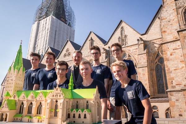 Gruppe von Jugendlichen in blauen T-Shirts steht hinter einem Modell des Paderborner Doms vor der echten Kathedrale