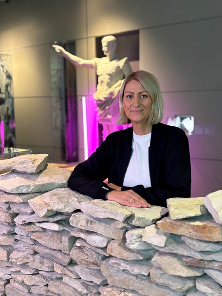Frau mit kurzen blonden Haaren und schwarzem Blazer lehnt an einer niedrigen Steinmauer in einem Museum mit einer antiken Statue im Hintergrund