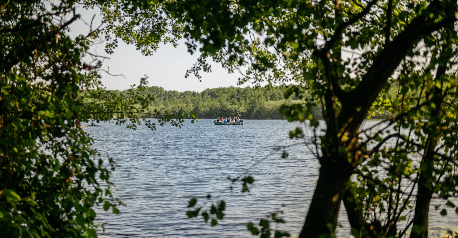 Blick durch Geäst auf ein Ruderboot in der Mitte eines Sees mit stiller Wasseroberfläche.