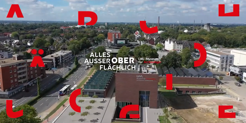 Eine Luftbildansicht vom Museumsgebäude, dazu der Titel "Alles außer oberflächlich!" und rote Buchstaben, die das Wort Archäologie ergeben.