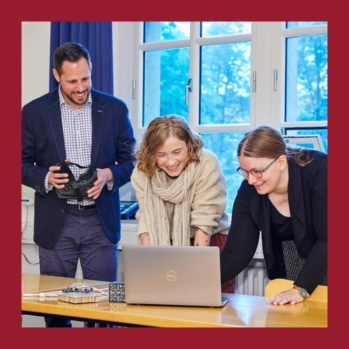 Drei Personen stehen an einem Tisch mit einem Laptop und VR-Brille vor einem Fenster. Foto eingerahmt von dunkelrotem Rahmen.