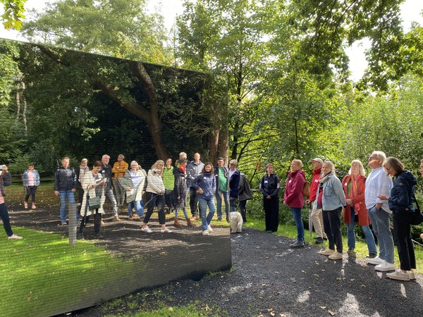 Gruppe von Menschen steht auf einem Weg im Park und blickt auf eine große spiegelnde Wand, die ihre Spiegelung zeigt