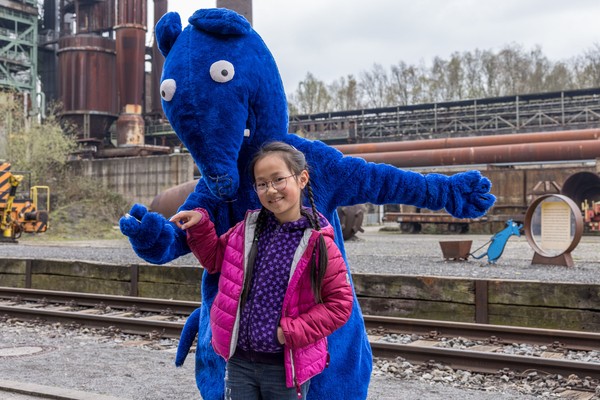 Kind mit pinker Jacke steht vor einer Person im blauen Rattenkostüm auf einem Gelände mit Bahngleisen und Industrieanlagen im Hintergrund.