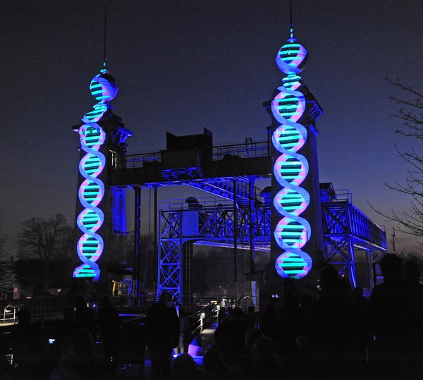 Eine dunkle Industrieanlage bei Nacht, auf zwei Türmen werden leuchtenden, spiralförmige Muster projiziert.