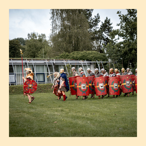 Gruppe von Menschen in römischer Legionärsausrüstung mit Schilden und Helmen marschiert auf einer Wiese vor einem modernen Gebäude. Eingefasst in gelbem Rahmen.
