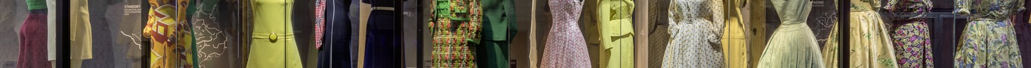 Lange Vitrine mit mehreren Damenkleidern in verschiedenen Mustern und Schnitten in einem Ausstellungsraum