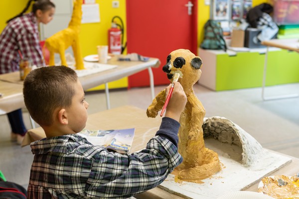 Kind mit kariertem Hemd bemalt eine gelbe Skulptur mit Pinsel in einem Raum mit roter Tür und weiteren Kunstobjekten.