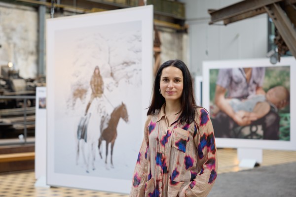 Eine Person mit langen schwarzen Haaren steht in einer Ausstellungshalle. Im Hintergrund hängen große Bilder.