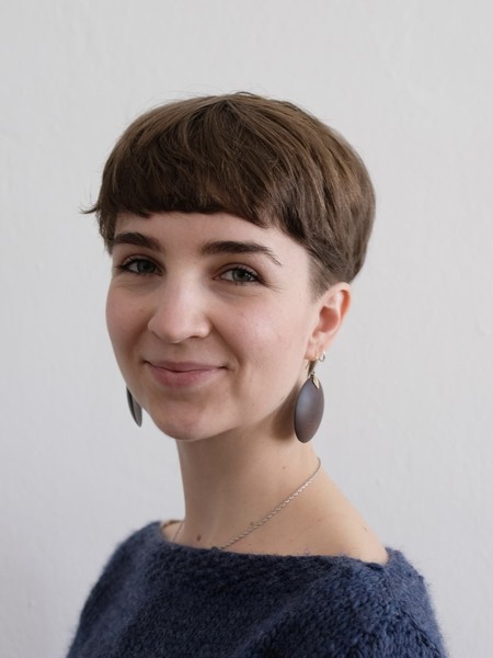 Frau mit kurzem braunem Haar, Ohrringen und blauem Pullover vor weißer Wand lächelnd