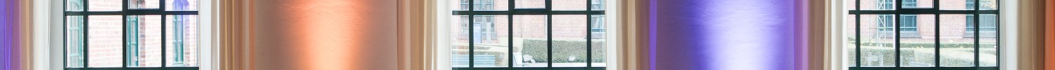 Publikum sitzt in einem Saal mit hohen, bogenförmigen Fenstern und farbiger Wandbeleuchtung