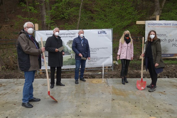Fünf Personen mit Schutzmasken stehen auf Betonboden, zwei halten rote Schaufeln, drei halten Dokumente, im Hintergrund Baupläne und Banner