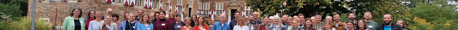 Große Gruppe von Menschen steht auf einem gepflasterten Platz vor einem historischen Backsteingebäude mit Baum und bewölktem Himmel.