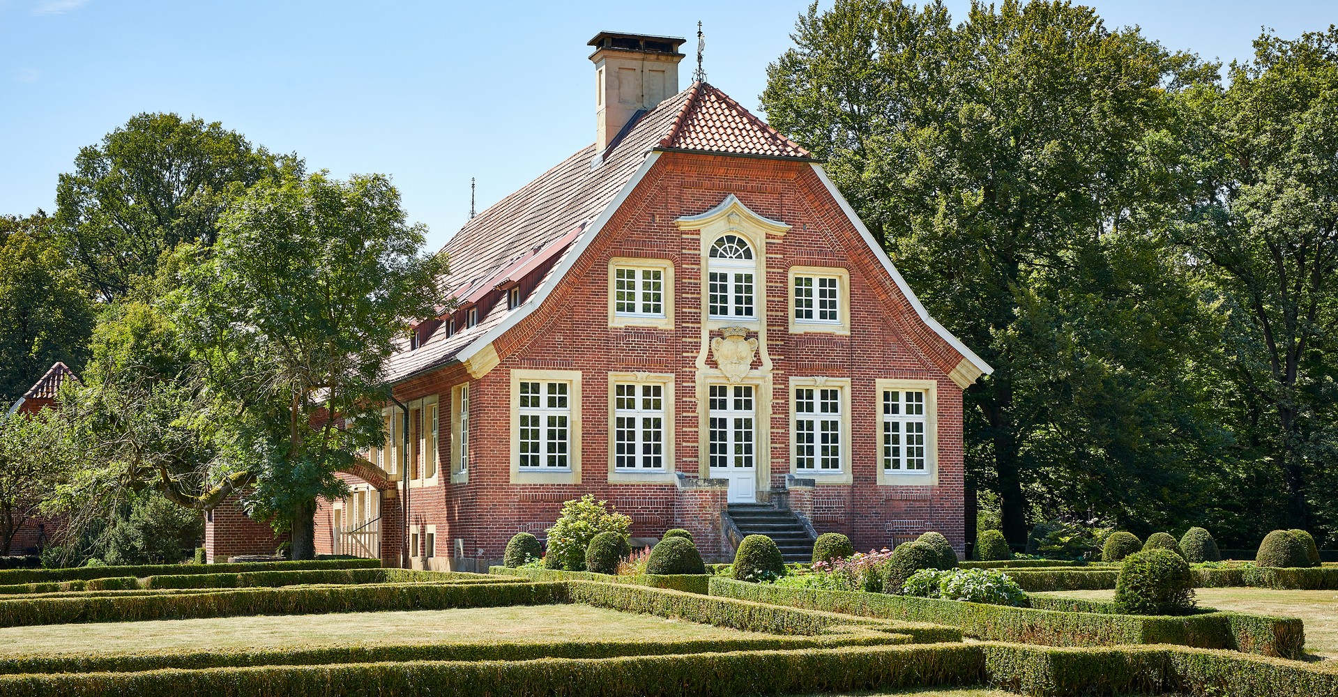 Ein Haus aus rötlichen Ziegelsteinen umgeben von einem grünem Garten.