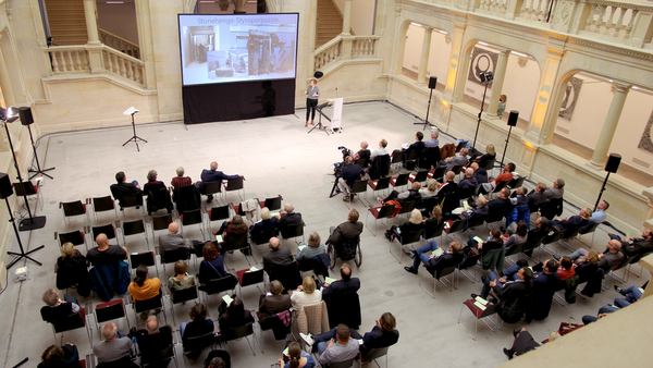 Großer Saal mit Publikum, das einem Vortrag vor einer Leinwand mit Präsentation folgt