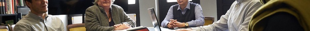 Fünf Personen sitzen an einem Konferenztisch, zwei nutzen Laptops und ein Tablet steht auf dem Tisch.