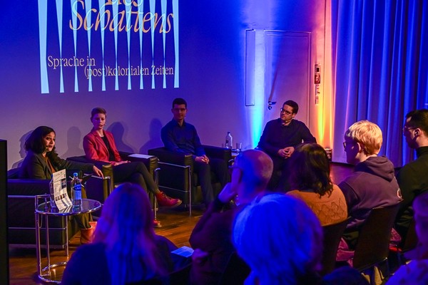 Podiumsdiskussion mit vier Personen auf Stühlen vor Publikum in einem Raum mit blauer Beleuchtung und  Schriftzug 'Sprache in (post)kolonialen Zeiten'