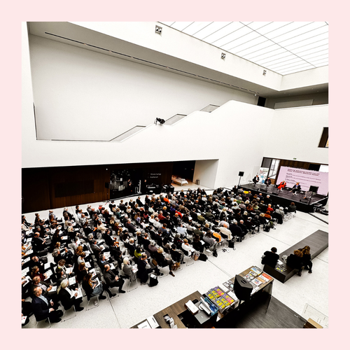 Großer Saal mit Publikum bei einer Konferenz vor einer Bühne mit drei sitzenden Personen und einer großen Leinwand. Foto eingerahmt von rosa Rahmen.